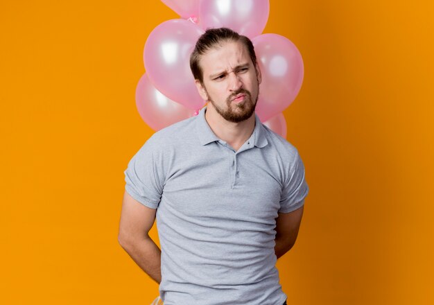 Young man celebrating birthday party holding bunch of balloons looking aside displeased and unhappy standing over orange wall