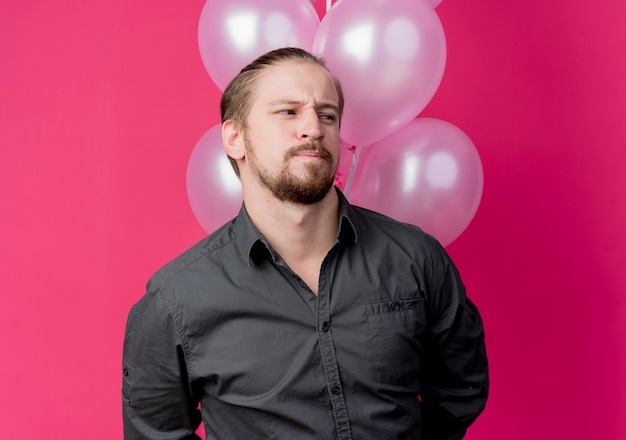 Giovane uomo che celebra la festa di compleanno tenendo un mazzo di palloncini guardando da parte perplesso in piedi sopra il muro rosa