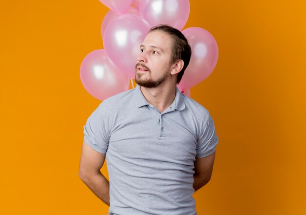 Young man celebrating birthday party holding bunch of balloons looking aside with pensive expression standing over orange wall