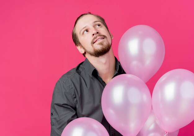 생일 파티를 축하하는 젊은 남자가 분홍색 벽 위에 서있는 생각에 잠겨있는 표정으로 올려다 보는 풍선의 무리를 들고
