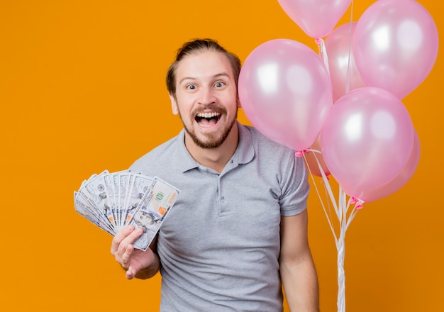 오렌지 벽 위에 현금 행복하고 흥분 서 보여주는 풍선의 무리를 들고 생일 파티를 축하하는 젊은 남자