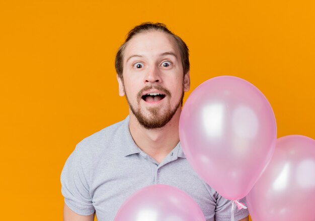 オレンジ色の壁の上に立って驚いた風船の束を保持している誕生日パーティーを祝う若い男