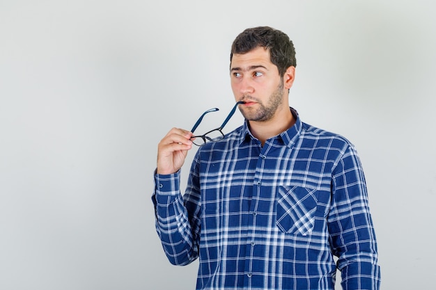 Free photo young man in checked shirt biting glasses and looking thoughtful