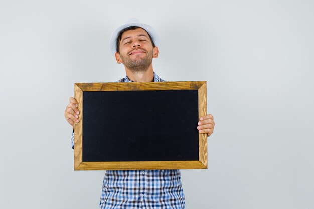 Young man in checked shirt, hat holding blackboard and looking peaceful