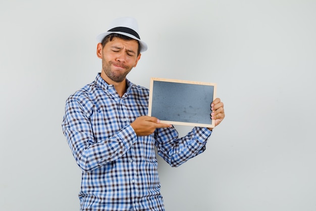 Молодой человек в клетчатой рубашке, шляпе держит пустую рамку с закрытыми глазами