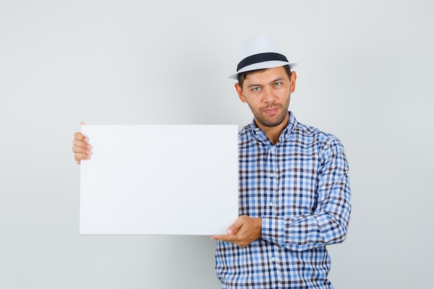 Young man in checked shirt, hat holding empty canvas and looking confident