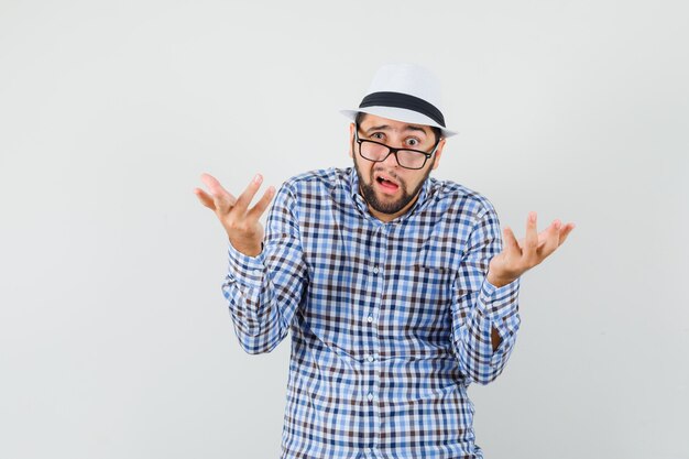 Young man in checked shirt, hat making asking question gesture and looking puzzled , front view.