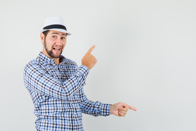 Young man in checked shirt, hat pointing fingers up and down and looking merry , front view.