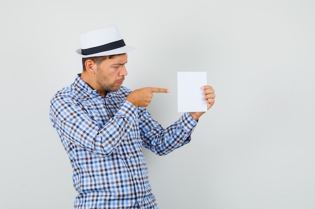 Young man in checked shirt, hat pointing at paper sheet and looking focused