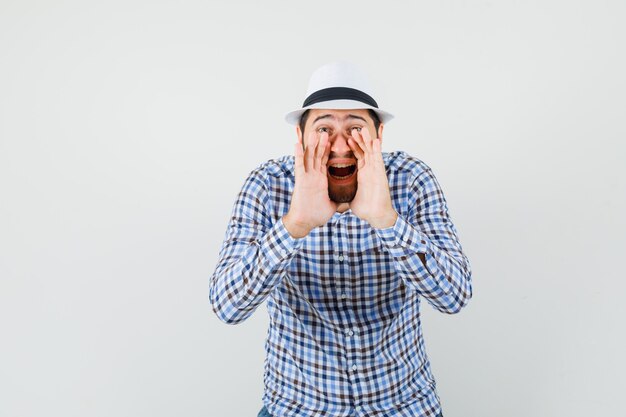 Young man in checked shirt, hat shouting or announcing something and looking excited , front view.