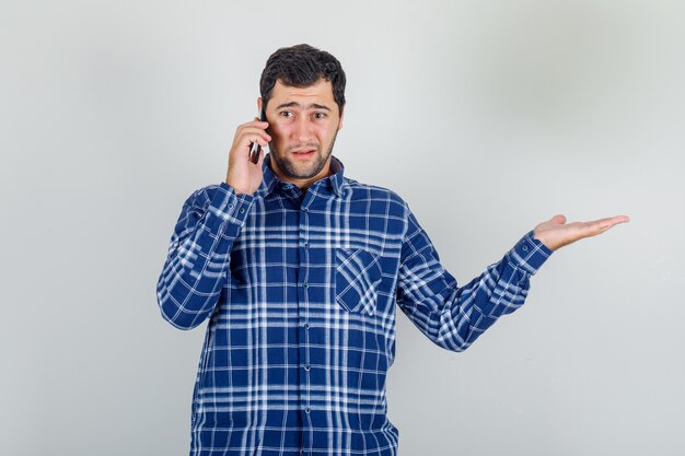 Молодой человек в клетчатой рубашке разговаривает по телефону жестом руки и выглядит разочарованным