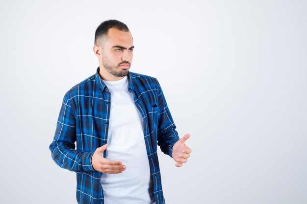 Young man in checked shirt and white t-shirt stretching hands as receiving something and looking annoyed