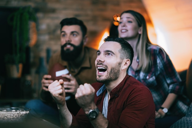 Young man cheering for his team while watching sports championship with friends