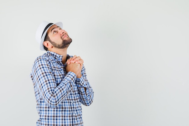 Young man clasping hands in praying gesture in shirt, hat and looking hopeful , front view.