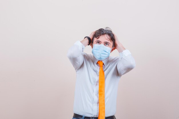 Young man clasping head in hands in shirt, tie, mask and looking scared