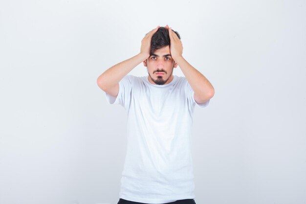 Young man clasping head while standing in t-shirt and looking forgetful