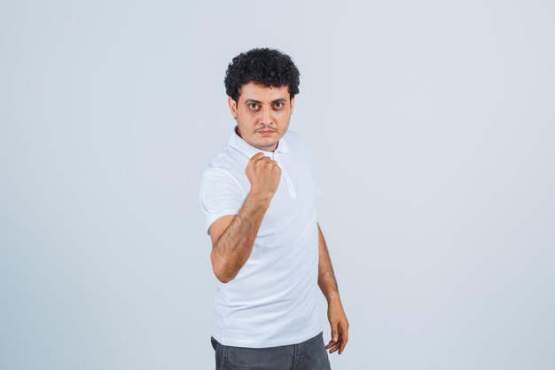 Молодой человек сжимает кулак в белой футболке и джинсах и выглядит серьезным, вид спереди.