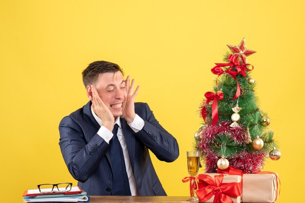 молодой человек, закрыв уши пальцами, сидит за столом возле рождественской елки и преподносит подарки на желтом