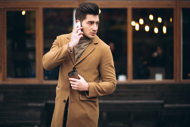 Young man in coat outside talking on the phone and drinking coffee