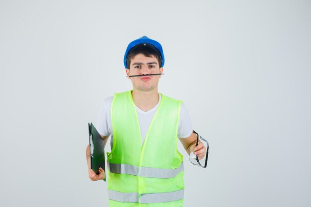 Молодой мальчик в строительной форме, держа в руках буфер обмена и защитные очки, держа ручку ртом и глядя серьезно, вид спереди.