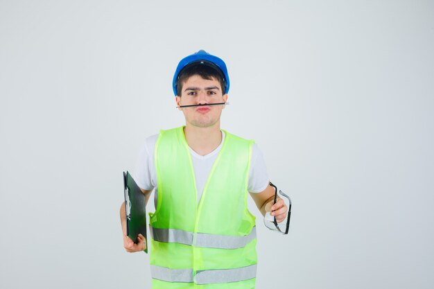 Young man in construction uniform holding clipboard and safety glasses in hands, holding pen with mouth and looking serious