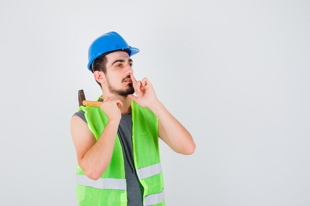 Young man in construction uniform raising axe over shoulder and showing silence gesture and looking focused