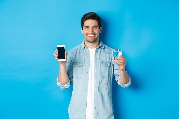若い男はスマートフォンアプリで水のバランスを制御し、モバイル画面アプリを表示し、青い背景の上に立って笑顔