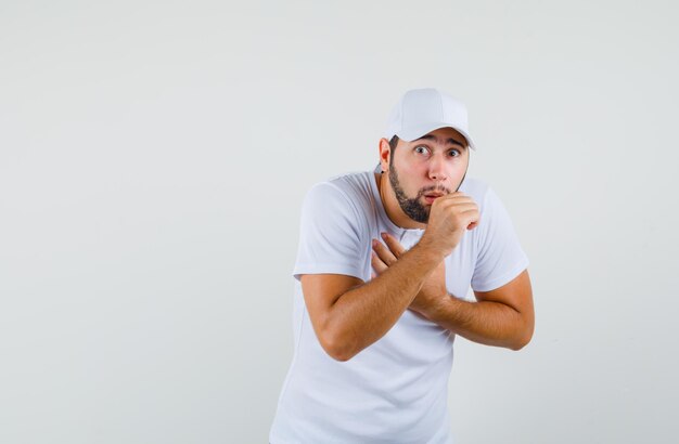Young man coughing in t-shirt,cap and looking sick , front view.
