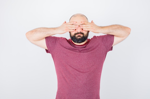 Free photo young man covering eyes with hands in pink t-shirt and looking serious. front view.