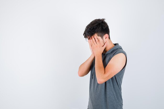 Young man covering face with hands in gray t-shirt and looking serious