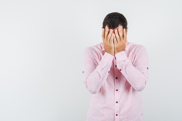 Young man covering face with hands in pink shirt and looking sad