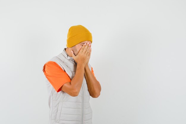Young man covering face with hands in t-shirt, jacket, hat and looking wistful. front view.