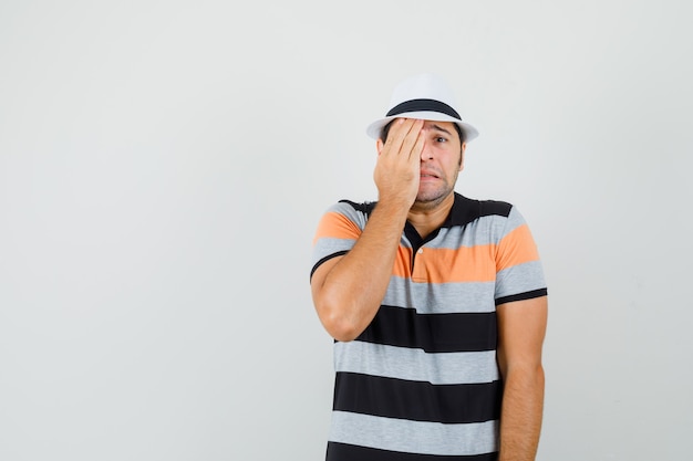 Young man covering face with his hand in striped t-shirt,hat and looking stressful space for text