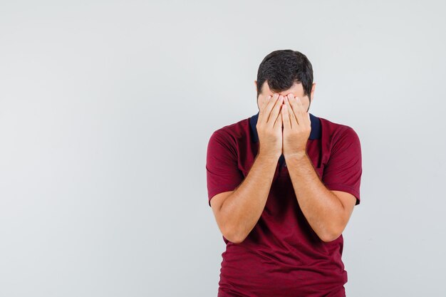 Young man covering his face with hands in red t-shirt and looking sad. front view.