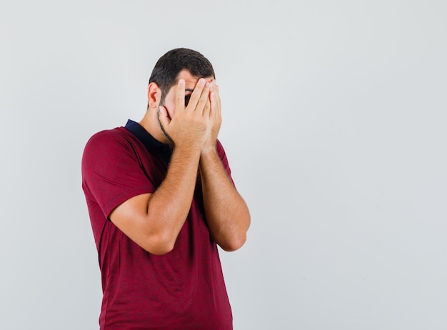 Young man covering his face with hands while looking through fingers in red t-shirt and looking scared , front view.