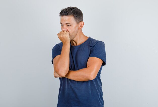 Young man covering mouth behind knuckle in dark blue t-shirt and looking sad, front view.