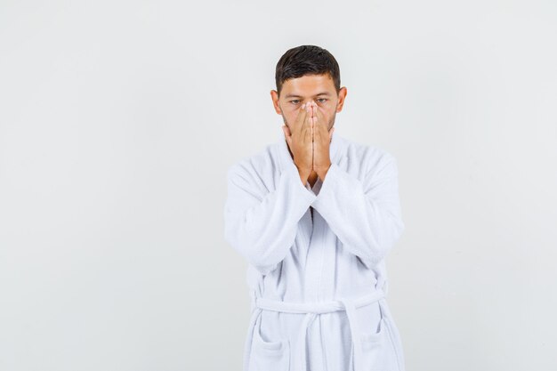 Young man covering mouth and nose in white bathrobe and looking pensive. front view.