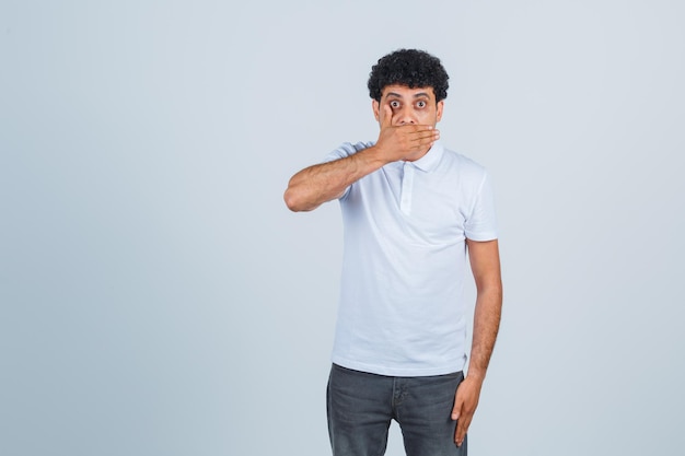 Young man covering mouth with hand in white t-shirt and jeans and looking surprised. front view.