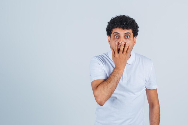 Young man covering mouth with hand in white t-shirt and jeans and looking surprised. front view.