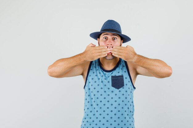 Young man covering mouth with hands in blue singlet, hat and looking scared