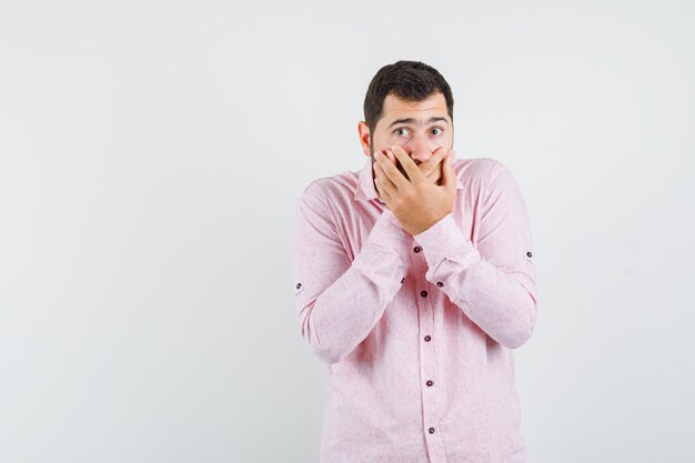 Young man covering mouth with hands in pink shirt and looking scared