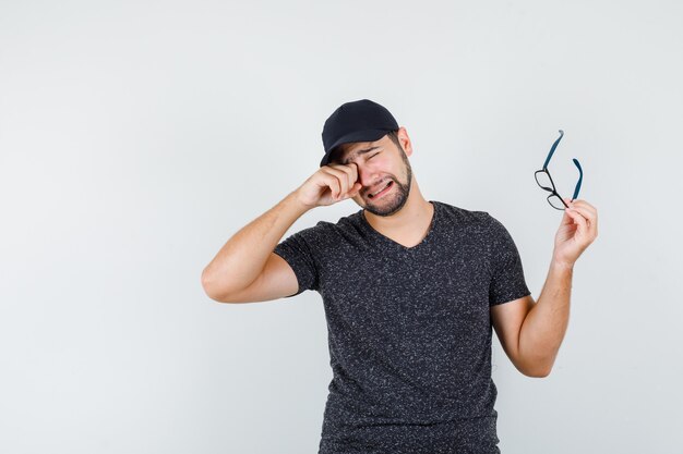 Young man crying while holding glasses in t-shirt and cap