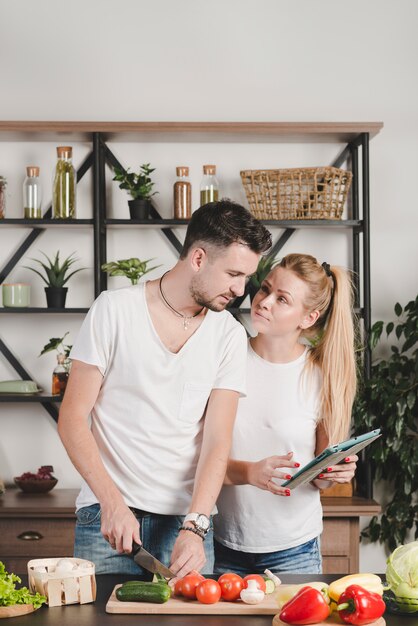 Young man cutting vegetables looking at digital tablet hold by her girlfriend