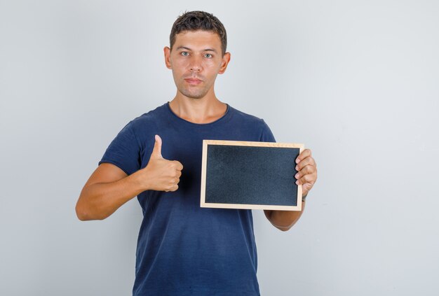 Young man in dark blue t-shirt holding blackboard and showing thumb up, front view.