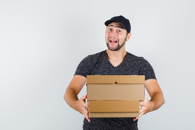 Young man delivering cardboard boxes in t-shirt and cap and looking glad