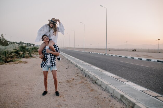 Young man in denim shorts holding his graceful girlfriend on shoulders standing near the highway. Adorable woman in vintage white blouse spending time with boyfriend and having fun on outdoor date