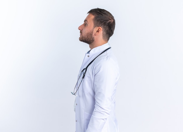 흰 코트를 입은 젊은 남자 의사와 목에 청진기를 들고 흰 벽 위에 옆으로 자신감 있게 서 있는 모습