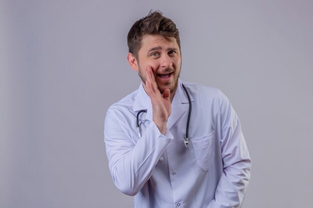 Young man doctor wearing white coat and stethoscope closing mouth whispering secret 