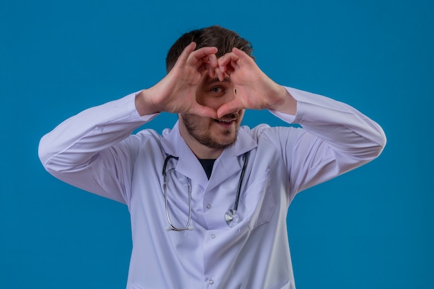 Young man doctor wearing white coat and stethoscope doing heart shape with hand and fingers smiling looking through sign over isolated blue background