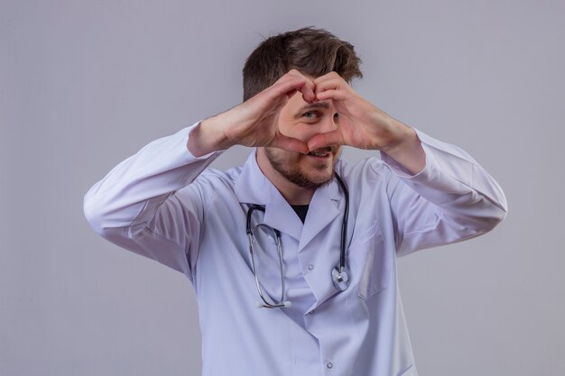 Young man doctor wearing white coat and stethoscope doing heart shape with hand and fingers smiling looking through sign